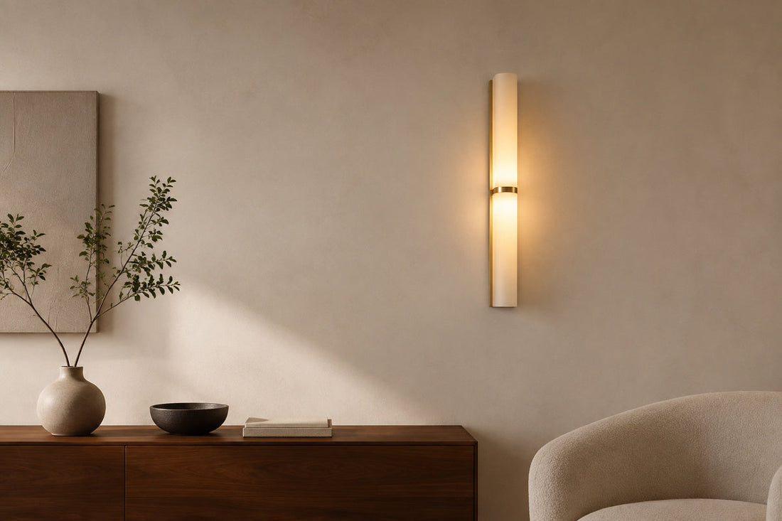 A lit brass and milky glass wall sconce above a dark wood console with a vase, bowl, books, and a cream chair.