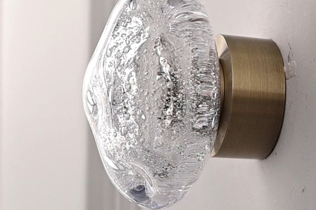 Exact Clear crystal-glass wall sconce illuminated on a pale wall, showing the textured glass globe and metal wall mount.