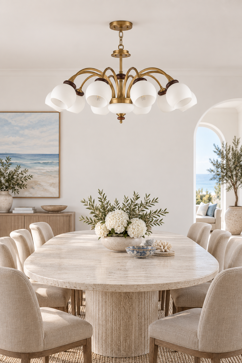 Arborenne ten-light chandelier centered above an oval stone table in a bright modern coastal dining room, fixture off