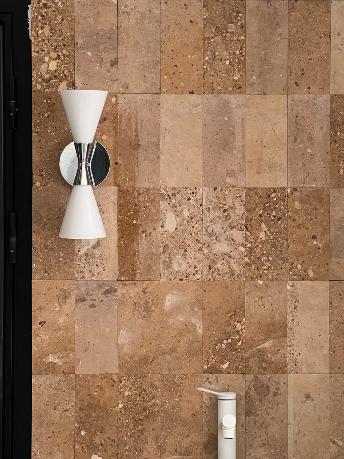 Polished chrome double-ended frosted glass wall sconce above a basin on a warm stone wall