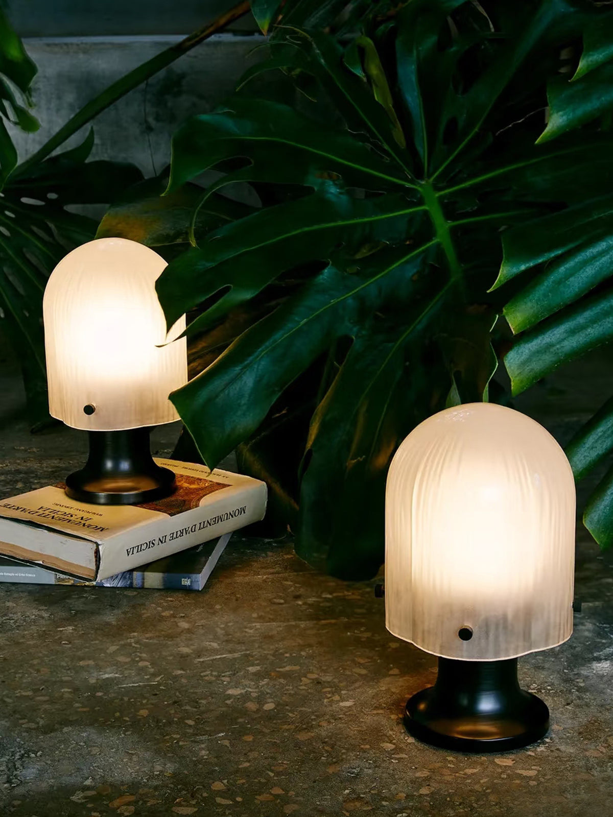 Two illuminated Mernoval streaked glass table lamps beside broad green leaves and books.