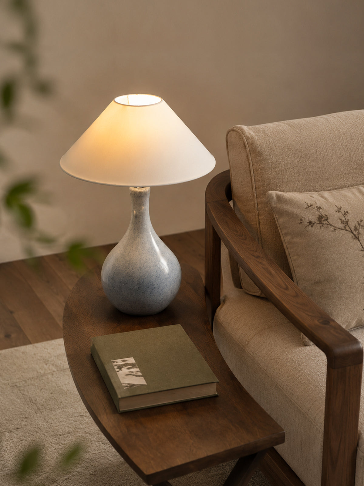 Lit blue-gray glazed ceramic table lamp beside a linen upholstered walnut armchair