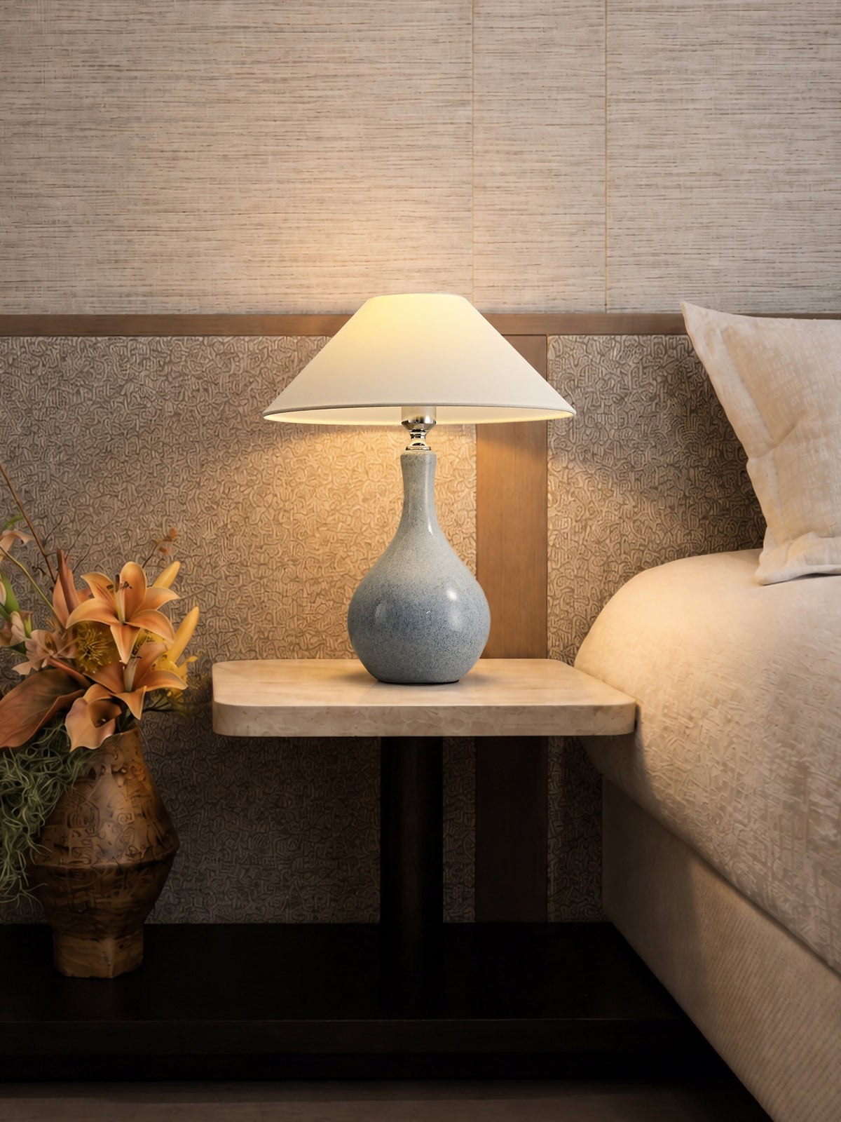 Lit blue-gray ceramic bedside lamp on a stone table with flowers and a textured headboard