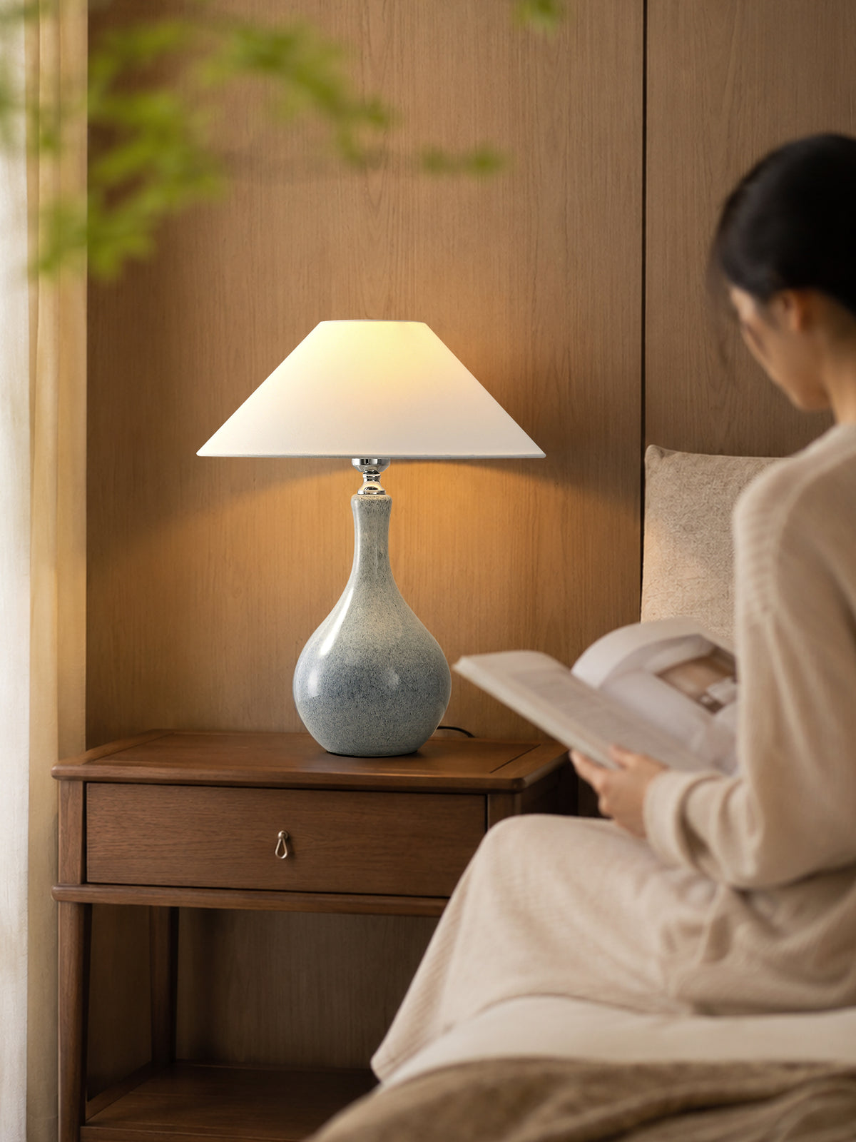 Lit blue-gray ceramic lamp on a walnut bedside table beside a seated reader