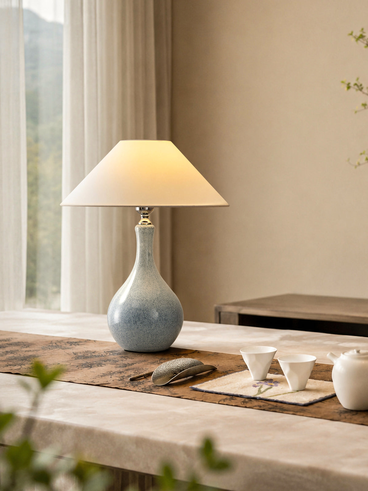 Lit blue-gray ceramic table lamp on a linen-covered tea table beside a bright window